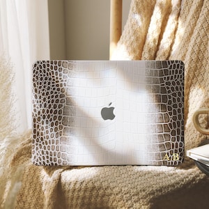 Puede incluir: Una funda para portátil con un diseño de piel de cocodrilo en blanco y marrón. El logotipo de Apple es visible. Las iniciales doradas "AB" están en la esquina inferior derecha. La funda está sobre una superficie texturizada.