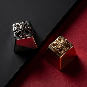 May include: Two mechanical keyboard keycaps, one in a polished silver finish and one in a polished gold finish. Each square keycap features a raised, ornate cross emblem on the top surface, characterised by four rounded lobes and a beaded centre.