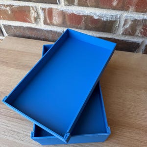 May include: Two bright blue rectangular trays are stacked at an angle against a brick wall and wooden surface. The trays have a simple, modern design with raised edges. The colour is a vibrant, solid blue.