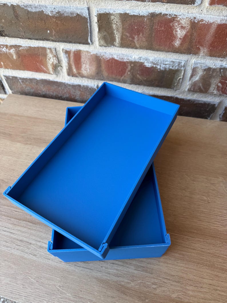 May include: Two bright blue rectangular trays are stacked at an angle against a brick wall and wooden surface. The trays have a simple, modern design with raised edges. The colour is a vibrant, solid blue.