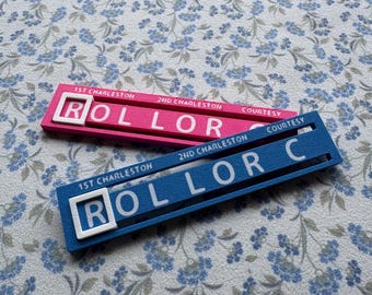 May include: Two rectangular game markers, one pink and one blue, featuring white raised lettering. Each marker displays the text 1ST CHARLESTON, 2ND CHARLESTON, COURTESY, and the word ROLLOR C with a white square frame around the letter R.