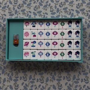 May include: A vintage Mahjong set in a teal case, featuring white tiles with various symbols, including flowers, lanterns, and directional markers. The set includes tiles marked "JOKER". A small bottle is also visible.