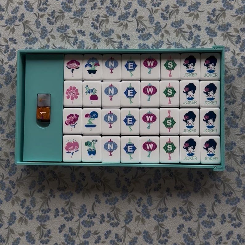 May include: A vintage Mahjong set in a teal case, featuring white tiles with various symbols, including flowers, lanterns, and directional markers. The set includes tiles marked "JOKER". A small bottle is also visible.
