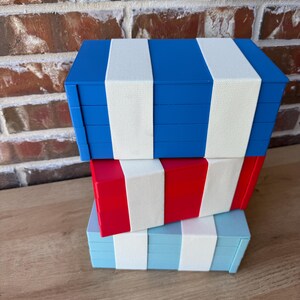 May include: Three decorative storage boxes in blue, red, and light blue, each with white stripes. The boxes are stacked on top of each other. The boxes are on a wooden surface. The boxes are likely for storing small items.