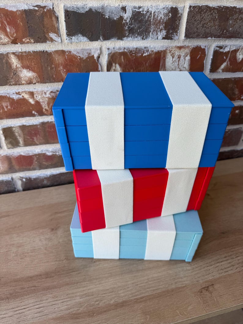 May include: Three decorative storage boxes in blue, red, and light blue, each with white stripes. The boxes are stacked on top of each other. The boxes are on a wooden surface. The boxes are likely for storing small items.