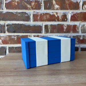 May include: A blue box with white bands, possibly for storage or display. The box is rectangular and has a simple, modern design. The background features a brick wall and a wooden surface.