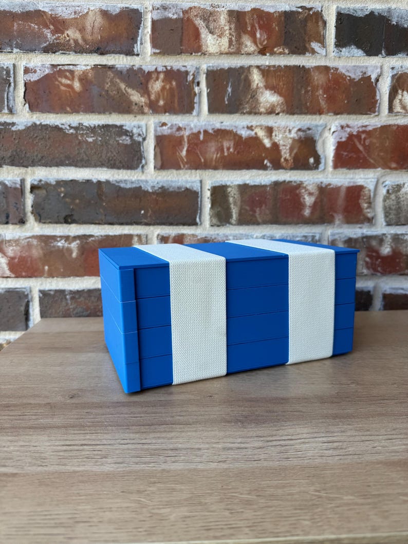 May include: A blue box with white bands, possibly for storage or display. The box is rectangular and has a simple, modern design. The background features a brick wall and a wooden surface.