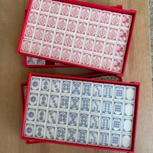 May include: Two red rectangular cases filled with Mahjong tiles. One case contains white tiles with red symbols, while the other has white tiles with blue symbols. The tiles are arranged neatly inside the cases.