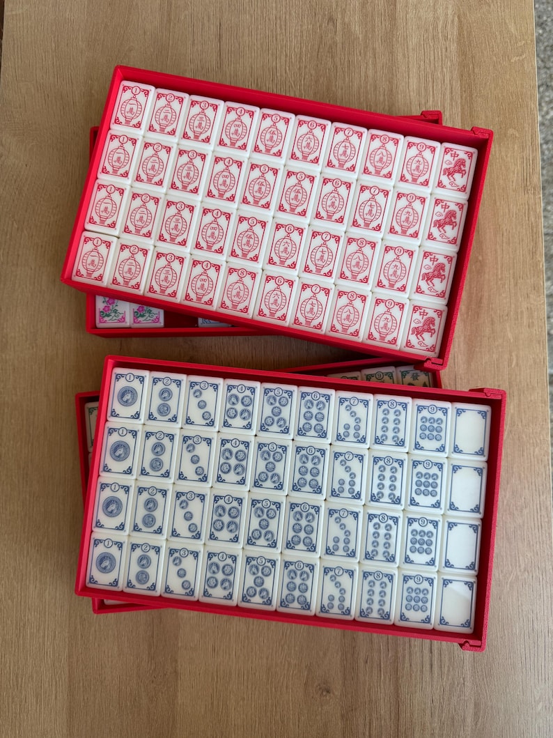 May include: Two red rectangular cases filled with Mahjong tiles. One case contains white tiles with red symbols, while the other has white tiles with blue symbols. The tiles are arranged neatly inside the cases.