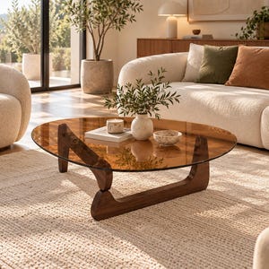 Peut inclure: Une table basse à profil bas dotée d'un plateau circulaire en verre teinté ambre transparent, reposant sur une base en bois brun foncé aux pieds incurvés et sculpturaux. Le plateau supporte un petit vase blanc moucheté, une bougie et un livre.
