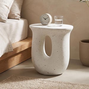 May include: A white speckled side table with a central oval cutout, featuring a smooth top surface. On the table sits a round analogue clock with a light-coloured frame and a clear glass of water. The table has a curved, sculptural base design.