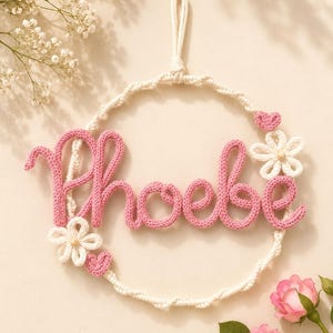 May include: A circular wall hanging featuring the name Phoebe in pink knitted yarn script. The name is centred within a cream-coloured twisted yarn hoop, decorated with two small white crocheted flowers and two tiny pink crocheted hearts.