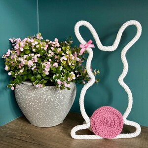 May include: A white knitted bunny decoration with a pink bow and a pink circular tail. A gray speckled pot with pink and green flowers is to the left of the bunny. The background is a teal colour.
