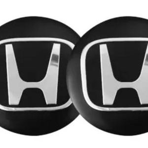 May include: A set of four circular wheel centre caps featuring a black background and a raised, metallic silver Honda logo in the centre. Each cap has a smooth, glossy finish with a distinct border surrounding the iconic H emblem design.