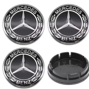Op de afbeelding: Vier Mercedes-Benz wieldopjes. Elke dop heeft een zilveren driepuntige ster op een zwarte achtergrond, omgeven door een zilveren ring met de woorden "MERCEDES BENZ". Eén dop wordt zonder het embleem getoond.