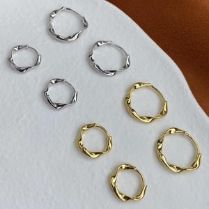 May include: A collection of seven small hoop earrings in silver and gold tones. Each earring features a distinct wavy, irregular texture along the circular band. The set includes four silver-colored hoops and three gold-colored hoops of varying sizes.