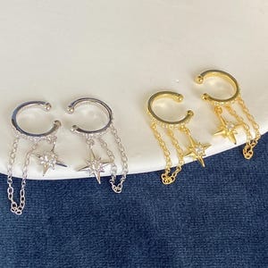 May include: Two pairs of ear cuffs, one silver-toned and one gold-toned, each featuring a circular band encrusted with small clear stones. Two delicate chains hang from each cuff, connecting to a central star-shaped charm with a single stone.