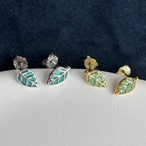 May include: Two pairs of leaf-shaped stud earrings. One pair is silver-toned with teal enamel inlay, and the other pair is gold-toned with light green enamel inlay. Each earring features a detailed vein pattern and a secure post-back closure.