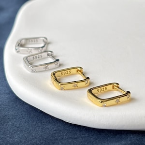 Könnte beinhalten: Zwei Paar rechteckige Creolen, ein Paar in Silber und ein Paar in Gold. Jeder Ohrring verfügt über kleine sternförmige Aussparungen mit eingelassenen klaren Steinen und die Gravur S925 auf dem inneren Bügel mit Klappverschluss.