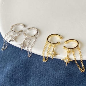 Può includere: Due paia di ear cuff, una color argento e una color oro, ciascuna con una fascia circolare incastonata con piccole pietre chiare. Ogni orecchino presenta due catenine sottili e un ciondolo a forma di stella con una pietra centrale.