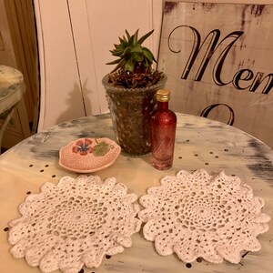 May include: Two white crocheted doilies on a white table with a small pink dish, a glass jar with a succulent plant, and a small glass bottle with a gold cap.