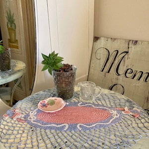 May include: A crocheted table runner with a pink and purple design. The runner is on a white table with a small plant and a glass pitcher.
