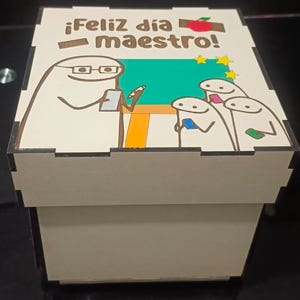 Può includere: Una scatola quadrata in legno con un coperchio che riporta il testo ¡Feliz día maestro! e una grafica colorata di un insegnante con gli occhiali davanti a una lavagna verde, mentre tre studenti guardano i loro telefoni, con una mela rossa e stelle gialle.