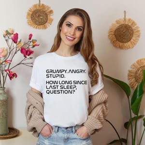May include: A white t-shirt with the text "GRUMPY. ANGRY. STUPID. HOW LONG SINCE LAST SLEEP, QUESTION?" in black. The shirt is paired with blue jeans and a beige cardigan. Decorative wall hangings and a vase of flowers are in the background.