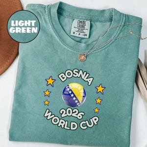 Pode incluir: T-shirt verde claro com um design da Copa do Mundo FIFA 2026 da B&oacute;snia. O design apresenta uma bola de futebol com as cores da bandeira da B&oacute;snia, rodeada por estrelas amarelas. A camiseta tem gola redonda e mangas curtas.