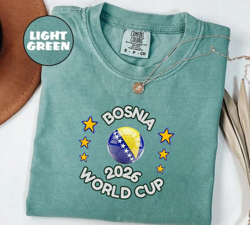 Pode incluir: T-shirt verde claro com um design da Copa do Mundo FIFA 2026 da B&oacute;snia. O design apresenta uma bola de futebol com as cores da bandeira da B&oacute;snia, rodeada por estrelas amarelas. A camiseta tem gola redonda e mangas curtas.