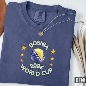 Pode incluir: Camiseta azul denim com um design da Copa do Mundo da B&oacute;snia 2026. O gr&aacute;fico apresenta uma bola de futebol com a bandeira b&oacute;snia, rodeada por estrelas. Um colar de prata com um pingente repousa sobre a camisa.
