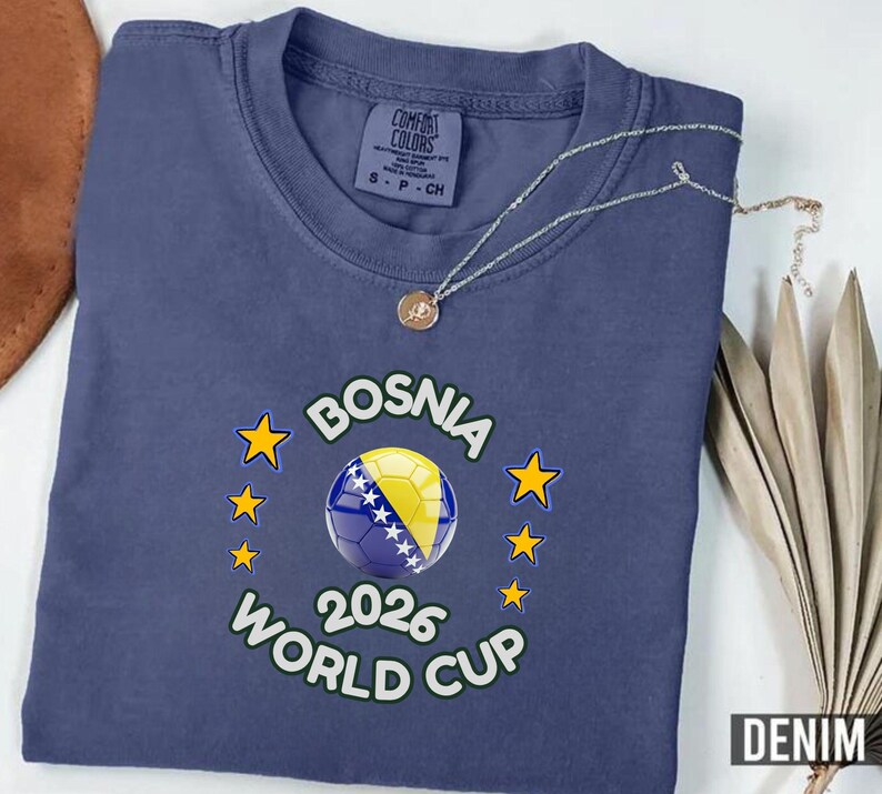 Pode incluir: Camiseta azul denim com um design da Copa do Mundo da B&oacute;snia 2026. O gr&aacute;fico apresenta uma bola de futebol com a bandeira b&oacute;snia, rodeada por estrelas. Um colar de prata com um pingente repousa sobre a camisa.