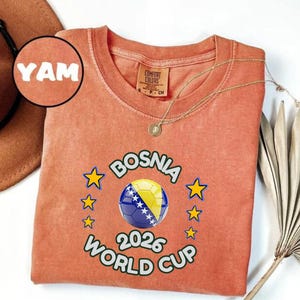 Pode incluir: Uma camiseta coral com o texto "BOSNIA WORLD CUP 2026" e um design de bola de futebol. A camisa tamb&eacute;m apresenta a palavra "YAM" e uma pequena etiqueta que diz "COMFORT COLORS". Um colar de ouro est&aacute; drapeado sobre a camisa.