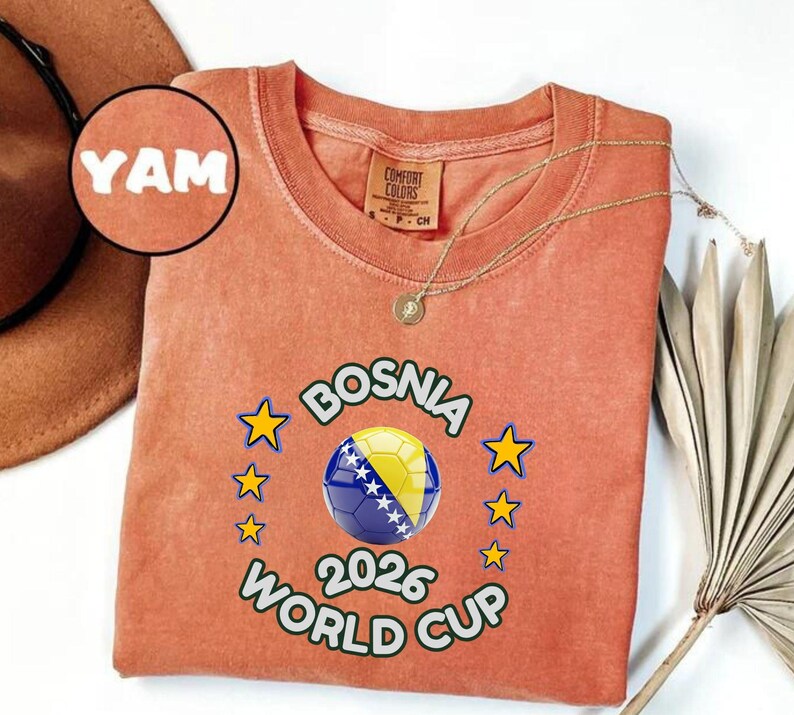 Pode incluir: Uma camiseta coral com o texto "BOSNIA WORLD CUP 2026" e um design de bola de futebol. A camisa tamb&eacute;m apresenta a palavra "YAM" e uma pequena etiqueta que diz "COMFORT COLORS". Um colar de ouro est&aacute; drapeado sobre a camisa.