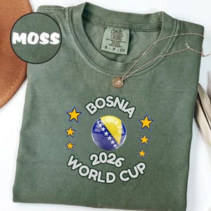Pode incluir: Uma t-shirt verde musgo com o texto "BOSNIA 2026 WORLD CUP" e um design de bola de futebol nas cores da bandeira b&oacute;snia. A t-shirt tem gola redonda e mangas curtas. Um colar de ouro est&aacute; sobre a t-shirt.