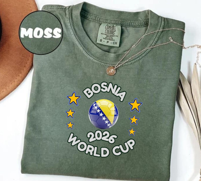 Pode incluir: Uma t-shirt verde musgo com o texto "BOSNIA 2026 WORLD CUP" e um design de bola de futebol nas cores da bandeira b&oacute;snia. A t-shirt tem gola redonda e mangas curtas. Um colar de ouro est&aacute; sobre a t-shirt.