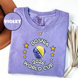 Pode incluir: Camiseta violeta com gola redonda. O design apresenta uma bola de futebol com a bandeira da B&oacute;snia, estrelas e o texto "BOSNIA 2026 WORLD CUP". Um colar dourado est&aacute; sobre a camiseta.