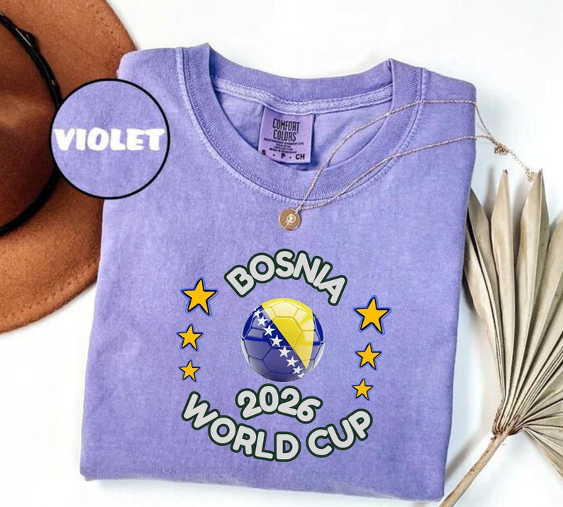 Pode incluir: Camiseta violeta com gola redonda. O design apresenta uma bola de futebol com a bandeira da B&oacute;snia, estrelas e o texto "BOSNIA 2026 WORLD CUP". Um colar dourado est&aacute; sobre a camiseta.