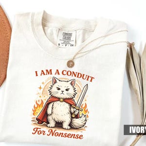 May include: Ivory-colored t-shirt with the text "I AM A CONDUIT For Nonsense" and a cartoon cat illustration. The cat is white with a red cape and a sword, surrounded by flames. A gold necklace is draped over the shirt.