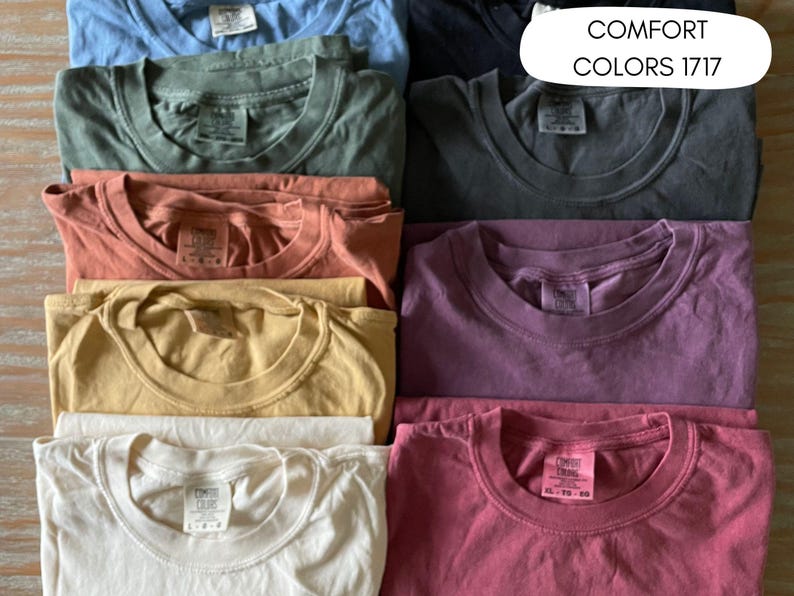Pode incluir: Uma cole&ccedil;&atilde;o de camisetas dobradas em v&aacute;rias cores, incluindo azul, verde, coral, amarelo, branco, cinza, roxo e rosa. As camisas t&ecirc;m gola redonda e s&atilde;o empilhadas. O texto "COMFORT COLORS 1717" &eacute; exibido em um oval branco.