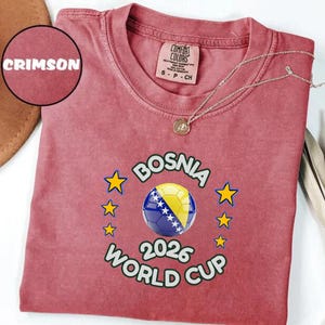 Pode incluir: Camiseta carmesim com um design da Copa do Mundo da B&oacute;snia 2026. O design apresenta uma bola de futebol com as cores da bandeira b&oacute;snia, rodeada por estrelas. A camisa tem gola redonda e mangas curtas.