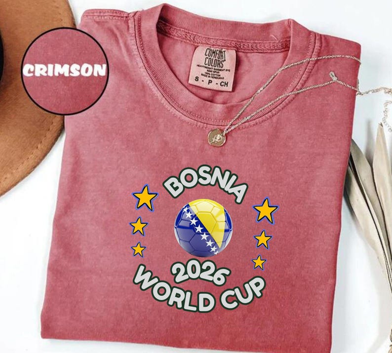 Pode incluir: Camiseta carmesim com um design da Copa do Mundo da B&oacute;snia 2026. O design apresenta uma bola de futebol com as cores da bandeira b&oacute;snia, rodeada por estrelas. A camisa tem gola redonda e mangas curtas.