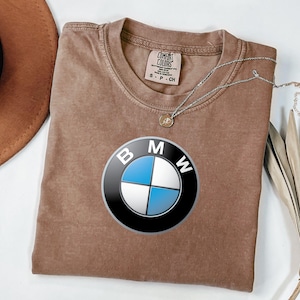 May include: A brown short-sleeved t-shirt featuring the BMW logo printed in the center. The shirt has a crew neckline and a Comfort Colors brand label on the inside collar with the text S P CH. A silver-toned necklace rests on the neckline.