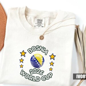Pode incluir: Camiseta cor marfim com um design da Copa do Mundo FIFA 2026 da B&oacute;snia. O design apresenta uma bola de futebol com as cores da bandeira b&oacute;snia, rodeada por estrelas e texto. A camiseta tem gola redonda e mangas curtas.