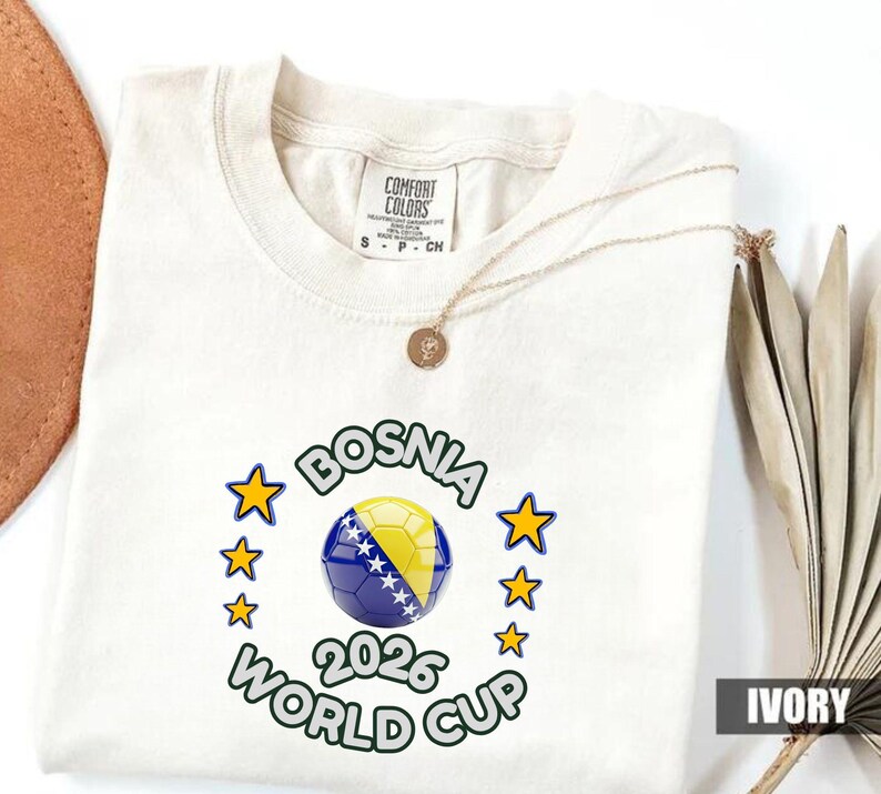 Pode incluir: Camiseta cor marfim com um design da Copa do Mundo FIFA 2026 da B&oacute;snia. O design apresenta uma bola de futebol com as cores da bandeira b&oacute;snia, rodeada por estrelas e texto. A camiseta tem gola redonda e mangas curtas.
