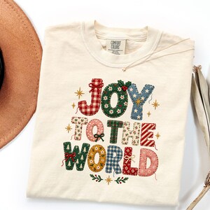 May include: A cream-colored short-sleeve t-shirt featuring a graphic print with the text JOY TO THE WORLD. The letters are designed with various patterns including red and white gingham, green floral, blue star, and plaid prints with small bows.