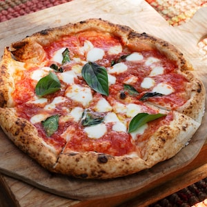 Pode incluir: Uma pizza recém-assada com uma crosta crocante, coberta com molho de tomate vermelho, queijo mussarela branco e folhas frescas de manjericão verde. A pizza é cortada e apresentada numa tábua de madeira.