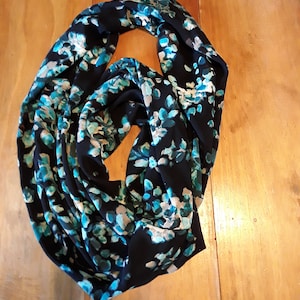 May include: A black infinity scarf with a floral pattern in shades of teal, light blue, and white. The scarf is draped on a wooden surface, showcasing its flowing fabric and design.