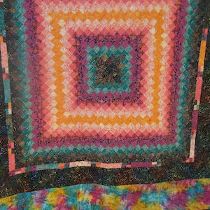 May include: A colorful handmade quilt with a square pattern. The quilt features a central square design in shades of pink, orange, and teal, surrounded by a black border and a tie-dye patterned edge. The quilt is made of fabric.