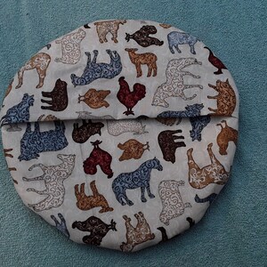May include: A round, cream-colored fabric cover with a repeating pattern of brown, blue, and red farm animals. The fabric is folded in half, showing the same pattern on both sides.